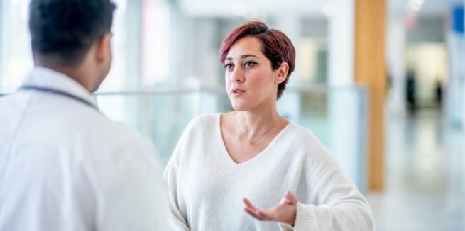 A woman of Middle Eastern ethnicity is meeting with her male physician. She is sitting at a table having a discussion with the doctor. The doctor has his back to the camera and the patient is holding out her hand as she tries to articulate her concern.